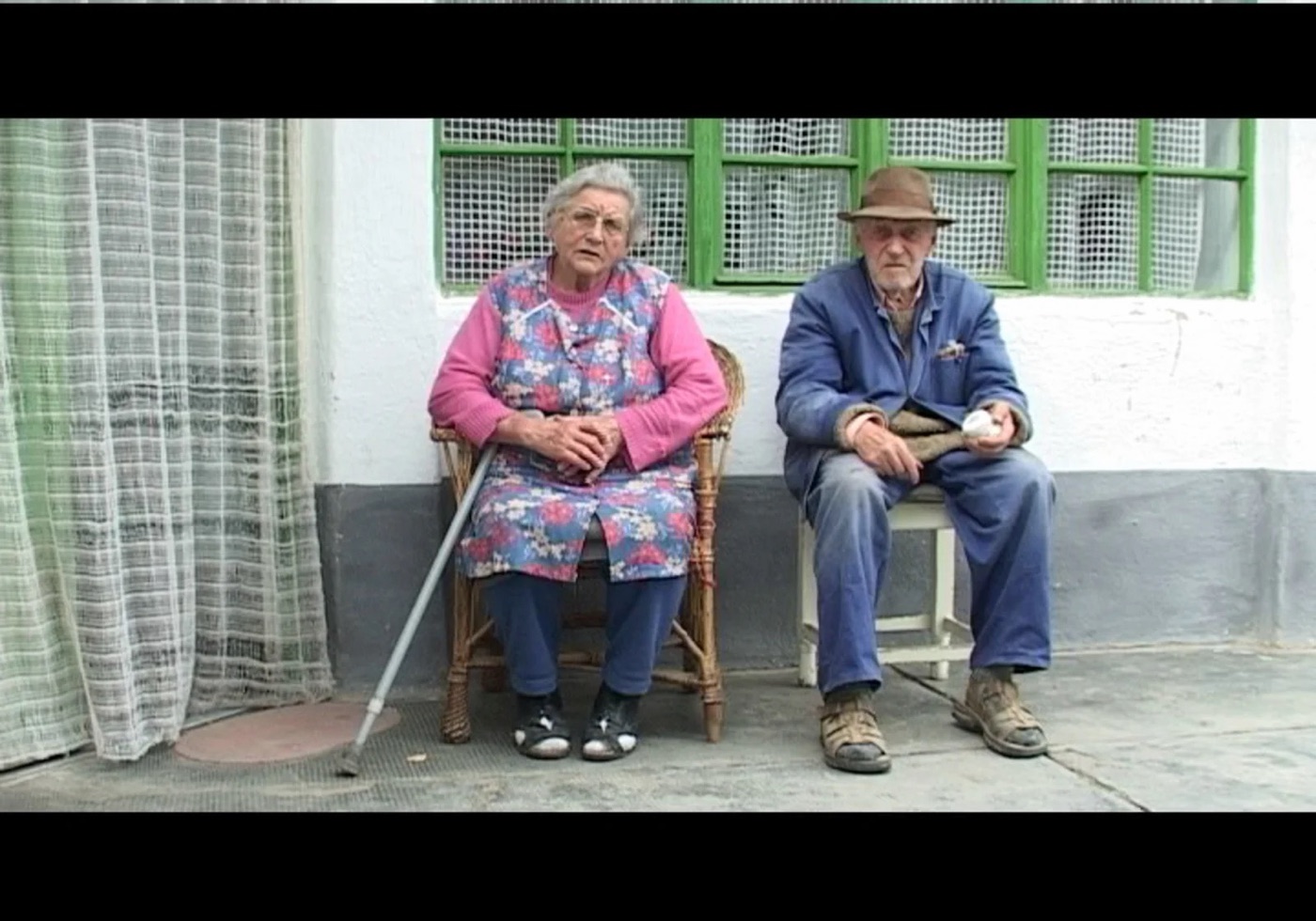 Still uit The Angelmakers: een ouder echtpaar op stoelen voor een wit huis.